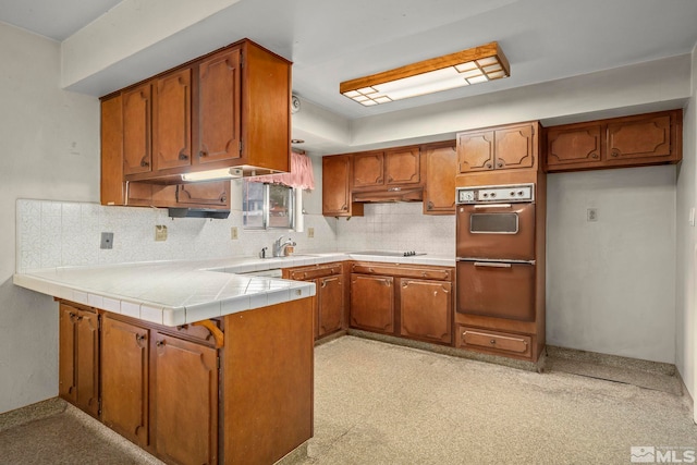 kitchen with brown cabinetry, a peninsula, black electric stovetop, under cabinet range hood, and a sink