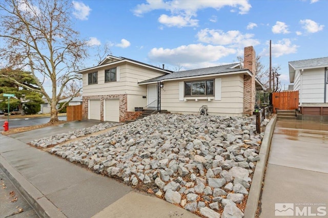 tri-level home featuring brick siding, a chimney, fence, a garage, and driveway