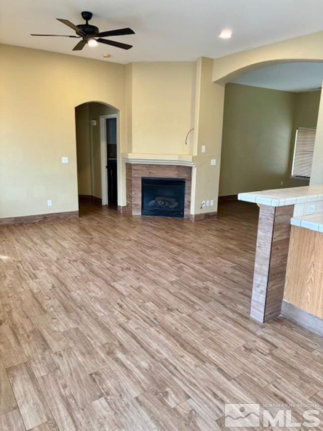 unfurnished living room featuring arched walkways, a fireplace, a ceiling fan, and light wood-style floors