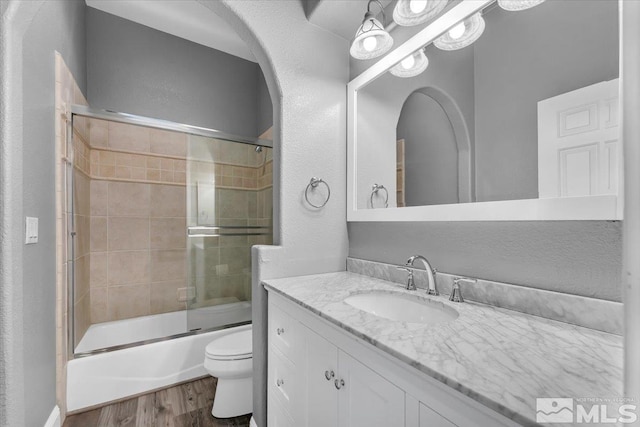 bathroom with bath / shower combo with glass door, a textured wall, toilet, wood finished floors, and vanity