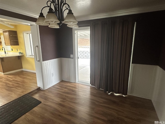 unfurnished dining area featuring a wainscoted wall, a sink, wood finished floors, an inviting chandelier, and crown molding