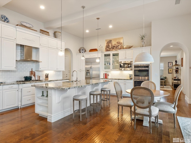 kitchen featuring arched walkways, built in appliances, wall chimney range hood, white cabinetry, and a sink