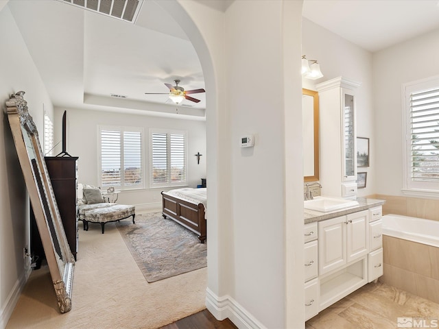 ensuite bathroom featuring a raised ceiling, ensuite bath, ceiling fan, vanity, and a bath