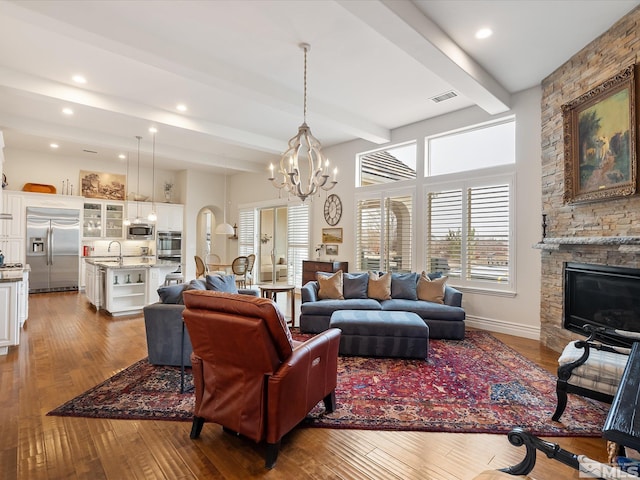 living room featuring baseboards, visible vents, hardwood / wood-style flooring, beamed ceiling, and a stone fireplace