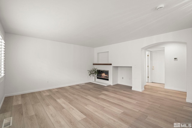 unfurnished living room with arched walkways, visible vents, baseboards, light wood finished floors, and a glass covered fireplace