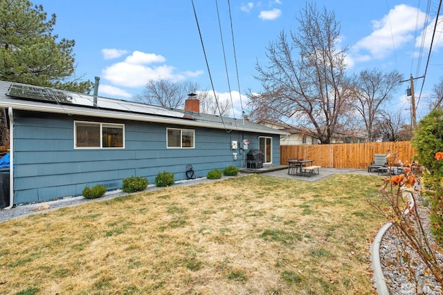 rear view of property with a patio, a fenced backyard, a lawn, roof mounted solar panels, and a chimney