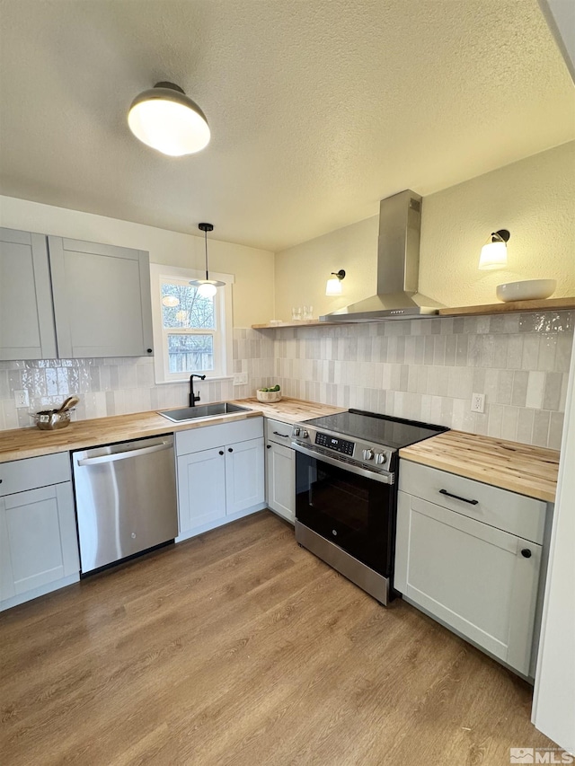 kitchen featuring wall chimney exhaust hood, butcher block countertops, appliances with stainless steel finishes, and a sink