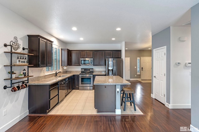 kitchen with dark brown cabinetry, stainless steel appliances, a sink, a center island, and light stone countertops