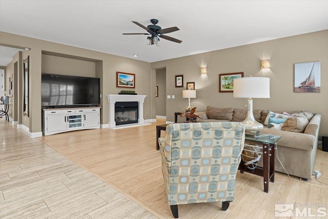 living room featuring a ceiling fan, light wood-type flooring, a glass covered fireplace, and baseboards