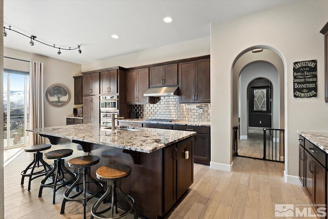 kitchen with dark brown cabinetry, arched walkways, appliances with stainless steel finishes, under cabinet range hood, and backsplash