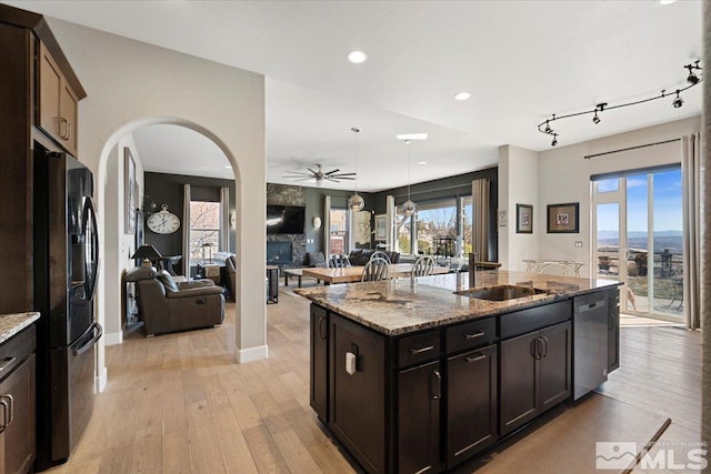 kitchen featuring stone counters, stainless steel dishwasher, black refrigerator with ice dispenser, light wood finished floors, and an island with sink