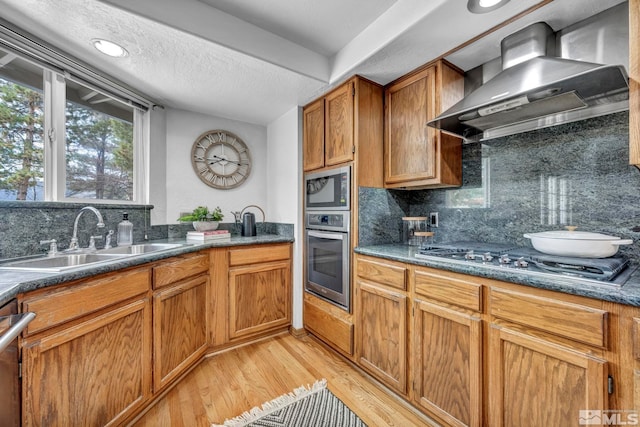 kitchen featuring light wood finished floors, appliances with stainless steel finishes, brown cabinetry, wall chimney exhaust hood, and a sink