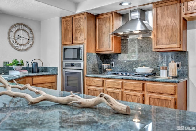 kitchen featuring wall chimney range hood, dark stone counters, decorative backsplash, brown cabinets, and stainless steel appliances