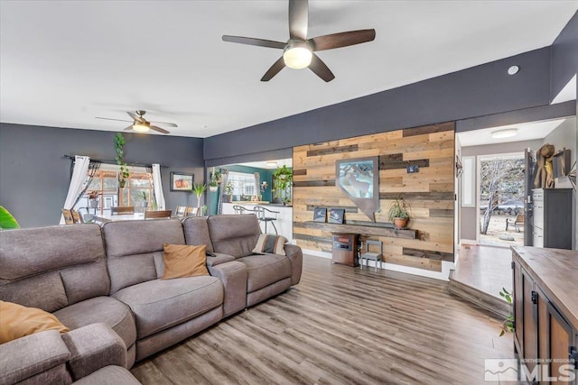 living room featuring wood finished floors, wooden walls, baseboards, ceiling fan, and an accent wall