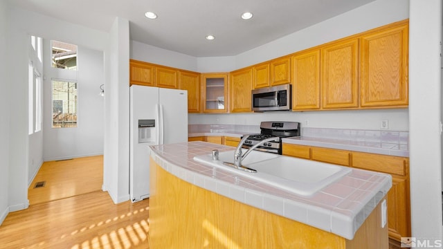 kitchen featuring glass insert cabinets, a center island with sink, tile countertops, light wood-style floors, and stainless steel appliances