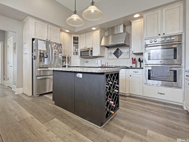 kitchen with a center island with sink, light wood finished floors, appliances with stainless steel finishes, wall chimney exhaust hood, and backsplash