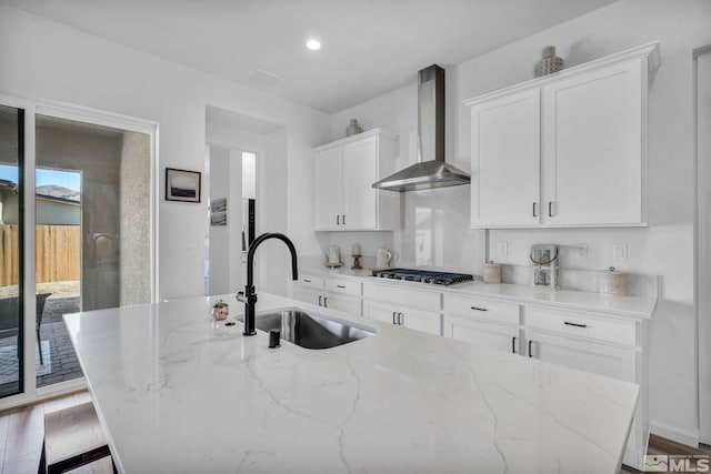 kitchen with light stone countertops, a kitchen island with sink, a sink, white cabinets, and wall chimney range hood