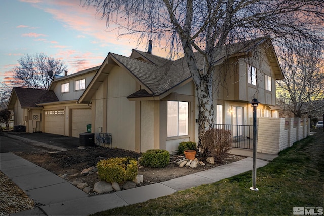 property exterior at dusk with fence, roof with shingles, an attached garage, stucco siding, and aphalt driveway