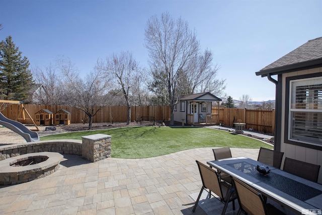 view of patio featuring a fenced backyard, a playground, a fire pit, an outdoor structure, and outdoor dining area