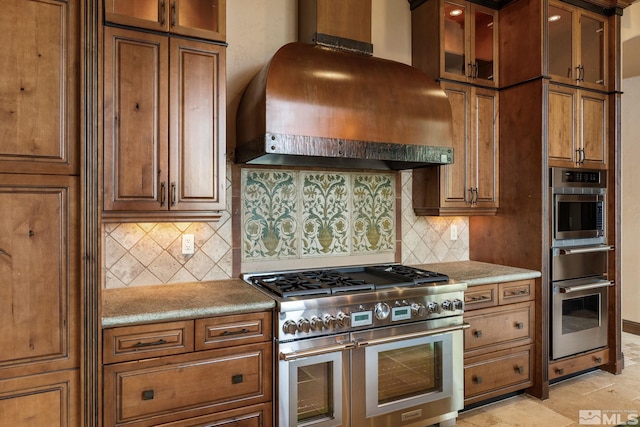 kitchen with glass insert cabinets, double oven range, decorative backsplash, brown cabinets, and custom exhaust hood