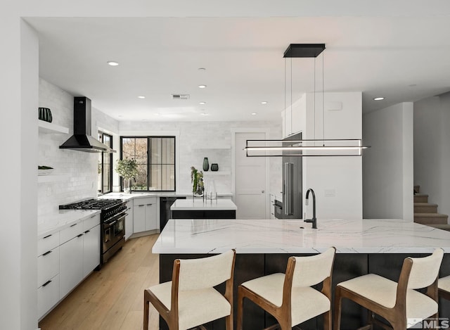 kitchen with range with two ovens, an island with sink, black dishwasher, wall chimney exhaust hood, and tasteful backsplash