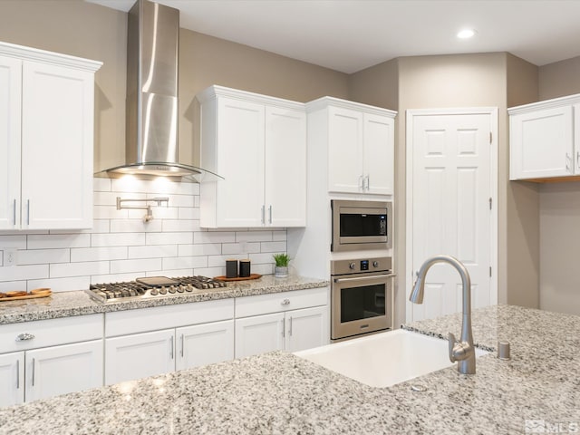 kitchen with a sink, decorative backsplash, white cabinets, appliances with stainless steel finishes, and wall chimney range hood