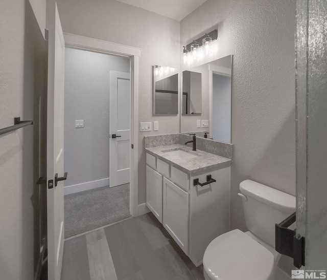 half bath with toilet, wood finished floors, baseboards, vanity, and a textured wall