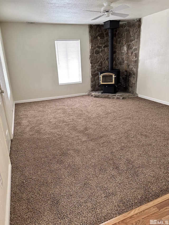 unfurnished living room with a ceiling fan, baseboards, carpet floors, a wood stove, and a textured ceiling
