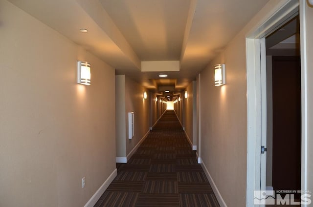 hall featuring dark colored carpet, baseboards, and a raised ceiling