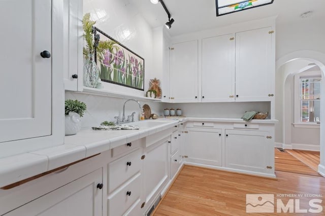 kitchen with arched walkways, a sink, white cabinetry, light wood finished floors, and tile countertops
