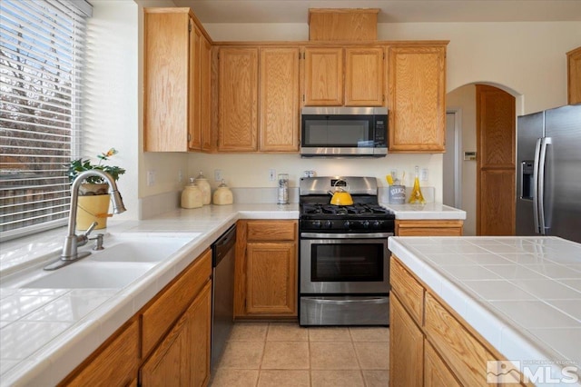 kitchen featuring light tile patterned floors, arched walkways, a sink, appliances with stainless steel finishes, and tile countertops