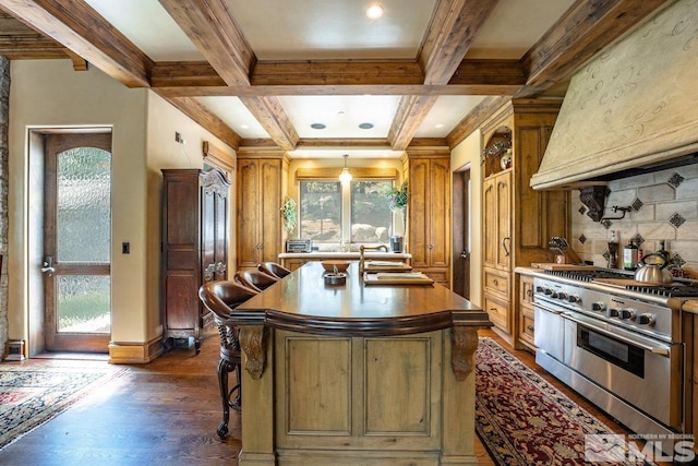 kitchen with coffered ceiling, stainless steel stove, beamed ceiling, wall chimney exhaust hood, and a center island with sink
