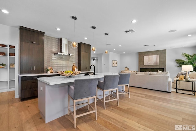 kitchen featuring a kitchen breakfast bar, light wood-style flooring, a kitchen island with sink, light countertops, and wall chimney exhaust hood