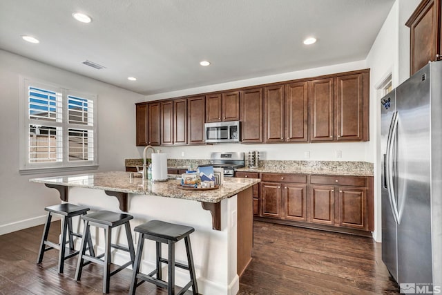 kitchen with dark wood-type flooring, a sink, light stone countertops, an island with sink, and appliances with stainless steel finishes