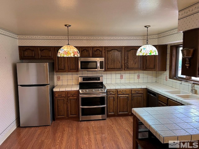 kitchen with wallpapered walls, appliances with stainless steel finishes, a sink, light wood-type flooring, and tile countertops