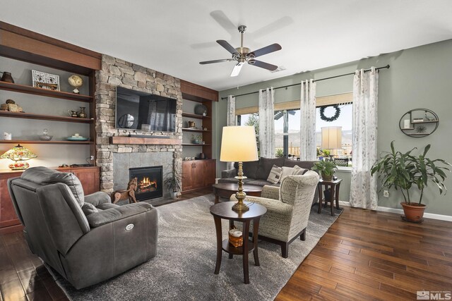 living area with wood-type flooring, a stone fireplace, built in features, a ceiling fan, and baseboards