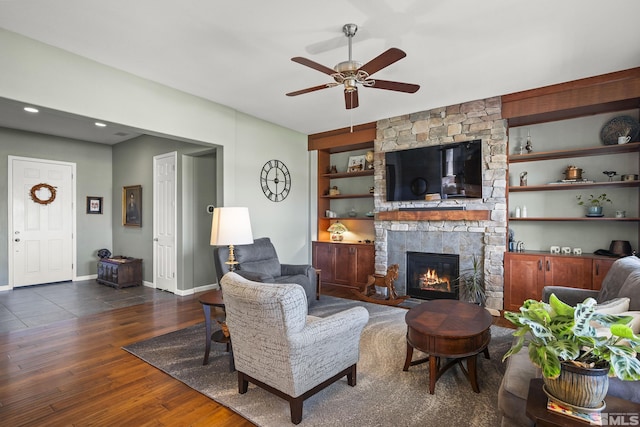 living room featuring a stone fireplace, baseboards, dark wood-style floors, built in shelves, and a ceiling fan