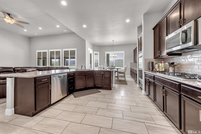 kitchen featuring dark brown cabinets, ceiling fan with notable chandelier, stainless steel appliances, open floor plan, and decorative backsplash