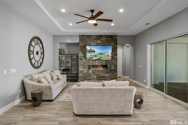 living room featuring ceiling fan, recessed lighting, a raised ceiling, a tiled fireplace, and light wood-style flooring