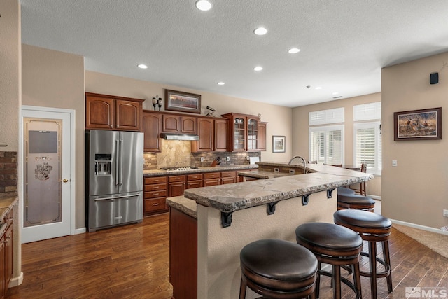 kitchen with a breakfast bar, dark wood finished floors, stainless steel appliances, under cabinet range hood, and glass insert cabinets