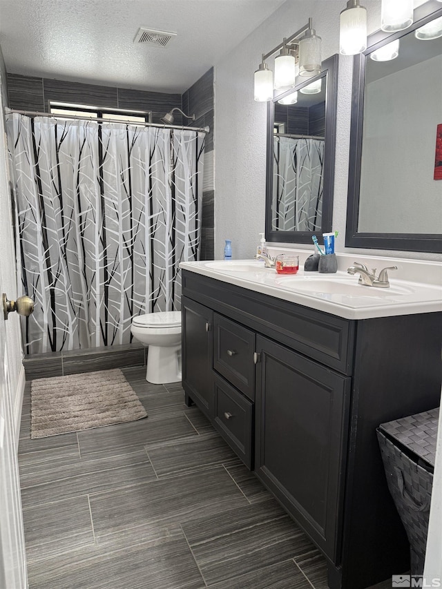 full bathroom with a textured wall, a sink, double vanity, toilet, and visible vents