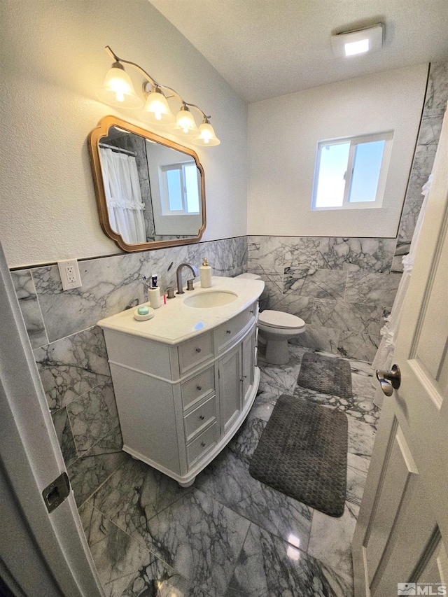 bathroom with tile walls, marble look tiles, vanity, a wainscoted wall, and toilet