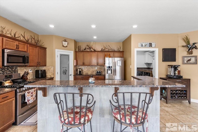 kitchen featuring a kitchen breakfast bar, brown cabinetry, a kitchen island, backsplash, and appliances with stainless steel finishes