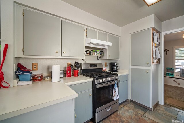 kitchen with stainless steel gas stove, under cabinet range hood, light countertops, stone finish floors, and gray cabinetry