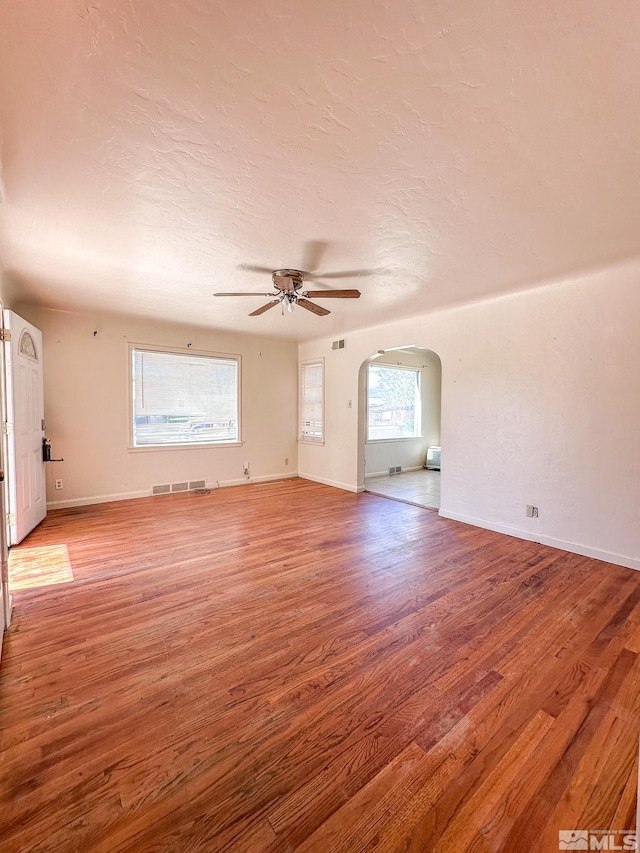 unfurnished living room with ceiling fan, arched walkways, wood finished floors, baseboards, and a textured ceiling