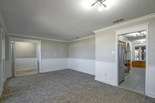 empty room with a textured ceiling, carpet flooring, visible vents, ornamental molding, and wainscoting