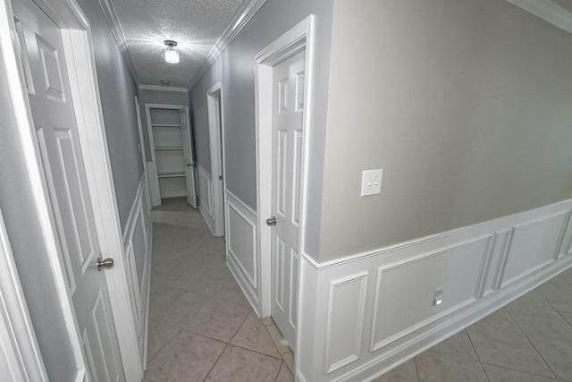 hall featuring a decorative wall, ornamental molding, wainscoting, light tile patterned flooring, and a textured ceiling