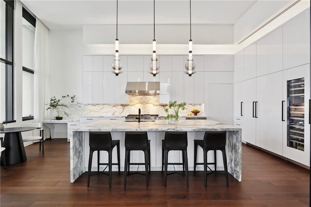 kitchen featuring extractor fan, light stone counters, decorative light fixtures, an island with sink, and white cabinets