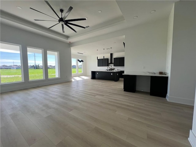 unfurnished living room with a tray ceiling, ceiling fan, sink, and light wood-type flooring