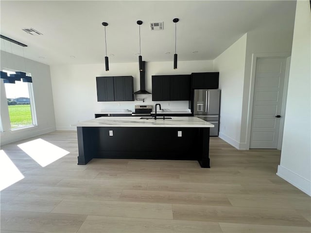 kitchen with hanging light fixtures, an island with sink, stainless steel appliances, and wall chimney range hood